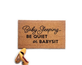 May include: A rectangular tan coir doormat with black text that reads Baby Sleeping... BE QUIET or BABYSIT. A pair of yellow slip-on shoes is positioned near the bottom left corner of the mat, which has a coarse, natural fiber texture.