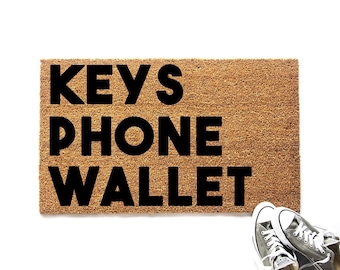 May include: A rectangular tan coir doormat featuring the black text KEYS PHONE WALLET printed in a bold sans-serif font. The mat has a coarse, fibrous texture and is positioned next to a pair of grey canvas sneakers with white laces and soles.