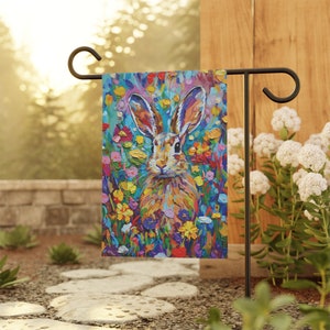 May include: A colorful garden flag featuring a painted image of a rabbit in a field of flowers. The rabbit is brown and white with long ears and a fluffy tail. The flowers are a variety of colors, including pink, purple, yellow, and blue.