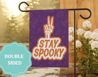 May include: A rectangular garden flag with a purple background featuring a repeating pattern of Halloween icons. A white skeleton hand makes a peace sign above the orange text "STAY SPOOKY" with a white outline. A teal label reads "DOUBLE SIDED".