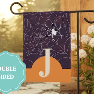 May include: A double-sided Halloween garden flag featuring a spider web design with a white spider and the letter "J". The flag has a purple background with an orange border and is displayed on a black metal stand.
