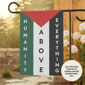May include: A rectangular garden flag featuring a red triangle at the top, with vertical green, white, and black stripes. The text "HUMANITY ABOVE EVERYTHING" is printed in white capital letters. A circular white badge states 100% PROCEEDS DONATED.