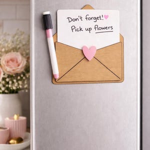 May include: A wooden envelope-shaped memo board with a white note that reads "Don't forget! Pick up flowers" and a pink heart. A black and pink marker is attached to the side. The memo board is on a silver refrigerator.