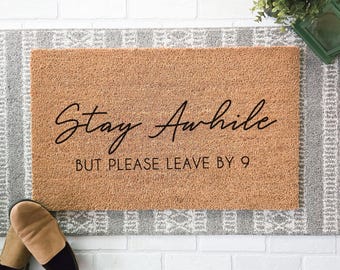 Peut inclure: Un paillasson rectangulaire en fibre de coco beige avec un texte noir indiquant Stay Awhile dans une police cursive, suivi du texte BUT PLEASE LEAVE BY 9 dans une police sans empattement. Le tapis a une texture fibreuse et grossière.