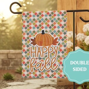May include: A rectangular garden flag featuring a repeating diamond pattern in orange, green, and white. The center displays an orange pumpkin illustration with brown leaves and the text "HAPPY fall" in white and orange lettering. A teal label reads "DOUBLE SIDED".