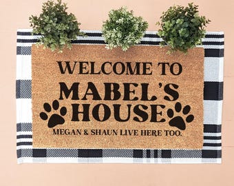 May include: A rectangular tan coir doormat layered over a black and white buffalo plaid rug. The doormat features black text reading WELCOME TO MABEL'S HOUSE and MEGAN & SHAUN LIVE HERE TOO, with two black paw prints flanking the word HOUSE.