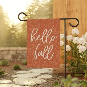 May include: A rectangular garden flag with the words "hello fall" in white script. The flag has a rust-colored background with a pattern of leaves, acorns, and other fall-themed elements. The flag is displayed on a black metal stand.