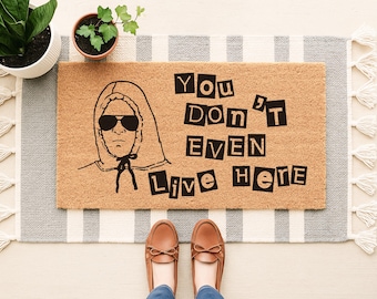 May include: A rectangular tan coir doormat featuring a black line-art illustration of a person wearing a hooded sweatshirt and sunglasses, with the black text "YOU DON'T EVEN LIVE HERE" printed in a bold, blocky font to the right of the illustration.