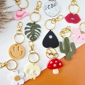 May include: A collection of acrylic keychains with gold-colored hardware. The keychains feature various designs, including a cowboy hat, a disco ball, lips, a bow, a smiley face, a monstera leaf, a planchette, a cactus, a mushroom, a skull, and a daisy.