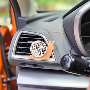 May include: A car air freshener clip shaped like a snail with a tan body and a white disco ball shell. The snail is attached to the horizontal slats of a black vehicle air vent. The disco ball shell features a grid pattern of white lines.