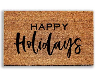 May include: A rectangular brown coir doormat featuring the black text HAPPY Holidays printed in the center. The mat has a coarse, fibrous texture typical of natural coconut husk fibers, providing a durable surface for entryways.