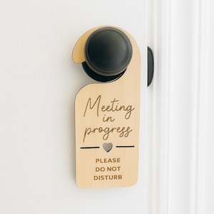 May include: A wooden door hanger with the words "Meeting in progress" and "Please do not disturb" engraved on it. The door hanger is hanging on a white door with a black doorknob.