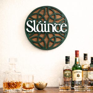 Könnte beinhalten: Ein rundes Wandschild mit einem dunkelgrünen, lasergeschnittenen Gittermuster auf einem dunklen Holzhintergrund. Der weiße Schriftzug "Sláinte" ist auf das Gitter aufgebracht. Das Schild hängt an einer Wand über einem Barbereich mit verschiedenen Flaschen.