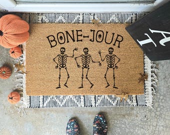 May include: A rectangular doormat with the words "BONE-JOUR" and three skeleton figures in black. The mat is on a patterned rug with orange pumpkins and autumn leaves. A black sign with white text is in the corner.