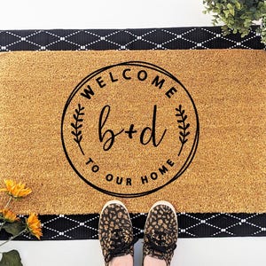 May include: A rectangular tan coir doormat featuring a black circular design with the text "WELCOME TO OUR HOME" and the letters "b+d" in the center. The mat is layered over a black rectangular rug with a white diamond pattern.