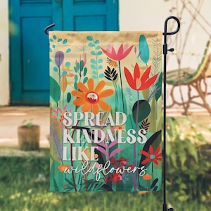 May include: A garden flag with a floral design and the words "SPREAD KINDNESS LIKE wildflowers" in white. The flag features orange, red, and pink flowers against a green and beige background. A black metal stand holds the flag.