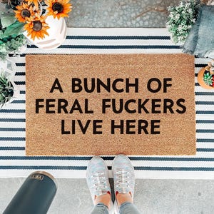May include: A rectangular tan coir doormat featuring the black text A BUNCH OF FERAL FUCKERS LIVE HERE, layered over a white and navy blue striped rectangular rug. The doormat has a coarse, natural fibre texture and bold, sans-serif lettering.