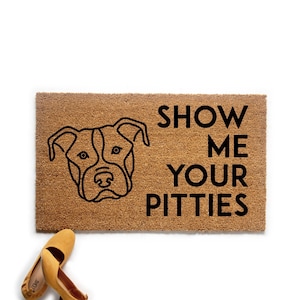 Peut inclure: Un paillasson rectangulaire en fibre de coco marron avec le contour noir d'un visage de chien et le texte "SHOW ME YOUR PITTIES" en noir. Une paire de ballerines jaunes est dans le coin inférieur gauche.