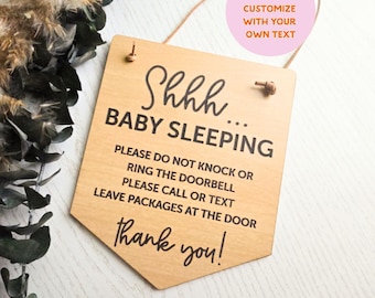 May include: A light-colored wooden sign with a pointed bottom edge, featuring black text that reads: Shhhh... BABY SLEEPING, PLEASE DO NOT KNOCK OR RING THE DOORBELL, PLEASE CALL OR TEXT, LEAVE PACKAGES AT THE DOOR, Thank you!. It hangs from a thin cord.