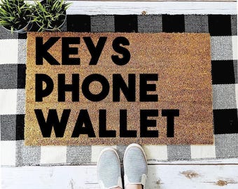May include: A rectangular tan coir doormat featuring the black text KEYS PHONE WALLET printed in a bold sans-serif font. The mat is positioned on a black and white buffalo check rug, with two small potted green plants visible at the top left.