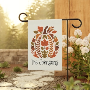 May include: A rectangular garden flag hanging on a black metal stand. The white flag features a central pumpkin illustration composed of various autumn-colored leaves and two acorns. The text "The Johnsons" is printed in black below the pumpkin.