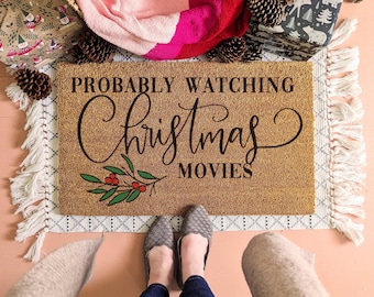 May include: A rectangular tan coir doormat featuring the black text "PROBABLY WATCHING Christmas MOVIES" in a mix of block and script fonts. A small illustration of a green holly branch with red berries is positioned below the text.