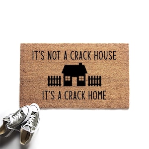 May include: A rectangular tan coir doormat featuring a black graphic of a house with a picket fence. The text printed in black reads: IT'S NOT A CRACK HOUSE IT'S A CRACK HOME. Two grey canvas trainers with white laces and rubber soles are positioned nearby.