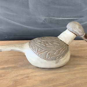 Peut inclure: Une sculpture d'oiseau en céramique blanche avec un motif texturé détaillé sur le dos. L'oiseau a un long bec fin et une tête ronde.