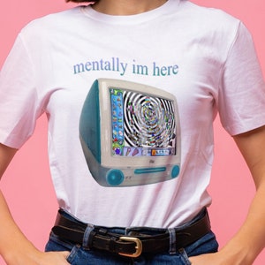 以下が含まれることがあります： 「mentally im here」というテキストと、渦巻くカラフルな画面を持つビンテージiMacコンピューターのグラフィックが描かれた白いTシャツ。