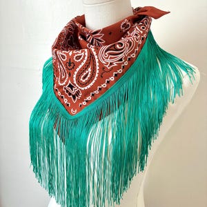 May include: A rust-colored bandana featuring a white and black paisley pattern, accented with a long, vibrant teal fringe trim along the bottom edge. The bandana is tied around the neck of a white mannequin, displaying the intricate print and fringe.