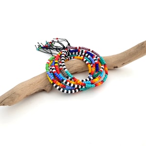 Può includere: Una collezione di braccialetti di perline colorate con vari motivi, tra cui strisce e colori pieni. I braccialetti sono disposti su un pezzo di legno trasportato dal mare su sfondo bianco. Le perline sono nei toni del rosso, blu, verde, giallo e nero.