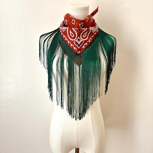 May include: A rust-colored bandana featuring a white paisley pattern, folded into a triangle and accented with long, dark green fringe that hangs down from the bottom edge, creating a layered appearance on a white mannequin torso.