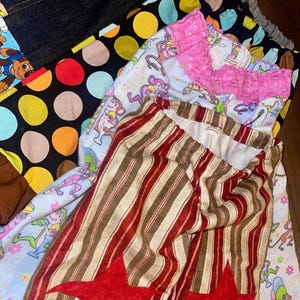 Puede incluir: Una colección de prendas que incluye tela de mezclilla oscura, un textil negro con lunares multicolores, una tela azul claro con gráficos de monos y helados con un borde de encaje rosa, y pantalones a rayas con aplicaciones de estrellas rojas.