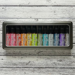 May include: A metal tin containing twelve small bottles of Distress Spritz ink. The bottles are arranged in a row and are labeled with the brand name and the color of the ink. The colors are a rainbow gradient from red to purple.