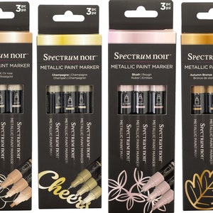 May include: Four sets of metallic paint markers in rose gold, champagne, blush, and autumn bronze. Each set contains three markers with different tip sizes. The packaging features the Spectrum Noir brand name and a decorative design.
