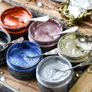 May include: Six small jars of metallic paint in various colors, including black, silver, gold, blue, copper, and pink. Each jar has a small spoon inside and is labeled "Jewel Effects Metallic Paint".