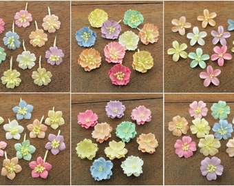 May include: Assortment of pastel colored paper flowers. The flowers are in various shades of pink, blue, yellow, and green. They are arranged in a grid pattern on a wooden surface.