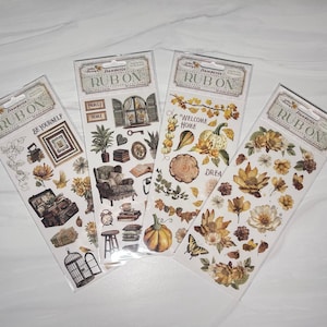 May include: Four packages of rub-on transfers with vintage-themed designs. The transfers feature images of flowers, pumpkins, furniture, and text like "BE YOURSELF" and "WELCOME HOME". The color palette includes shades of brown, yellow, and cream.