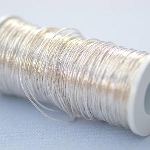 May include: A spool of silver metallic thread wound around a white plastic bobbin. The thread is shiny and has a subtle shimmer.