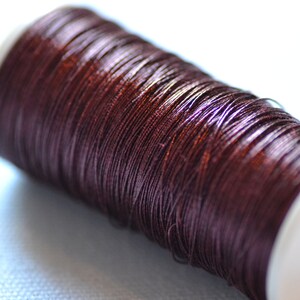 May include: A spool of dark brown metallic thread wound around a white plastic bobbin.