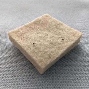 May include: A square, beige felt pad with a slightly textured surface. The pad has a diamond shape and appears to be made of a soft, natural material. Small dark specks are visible on the surface.