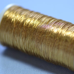 May include: Close-up of a spool of gold metallic thread. The thread is tightly wound around a white plastic spool. The metallic thread has a shiny, reflective surface, and the overall effect is one of luxury and craftsmanship. Ideal for embroidery and sewing projects.