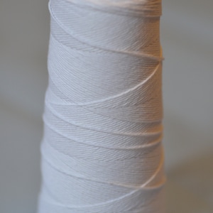 May include: A spool of white cotton thread wound around a black plastic bobbin.