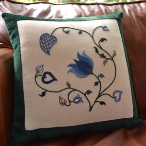 May include: A white pillow with a green border features an embroidered design of blue and green flowers and leaves.