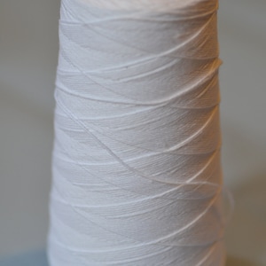 Könnte beinhalten: Eine weiße Garnspule, eng um einen konischen Kern gewickelt. Der Faden ist strahlend weiß und die Spule hat eine schwarze Kunststoffmitte. Der Faden ist sauber gewickelt und erzeugt eine strukturierte Oberfläche.