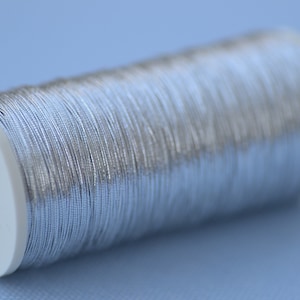 May include: A spool of silver metallic thread wound around a white plastic bobbin.