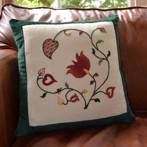 May include: A white cushion with a green border features an embroidered design of a red flower with yellow accents and green leaves. The design includes a small heart shape in the top left corner.