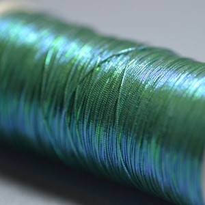 May include: Close-up of a spool of iridescent thread. The thread is a blend of green and blue hues, creating a shimmering effect. The spool has white sides and is ideal for sewing, embroidery, or other crafts.