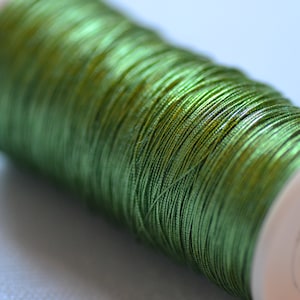 May include: A spool of green metallic thread wound around a white plastic bobbin.