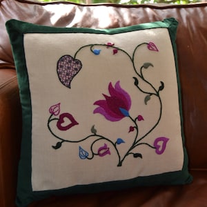 Könnte beinhalten: Ein weißes Kissen mit grünem Rand zeigt ein gesticktes Design aus rosa, lila und blauen Blumen mit grünen Stielen und Blättern. Die Blumen sind stilisiert und haben eine Herzform an der Basis.